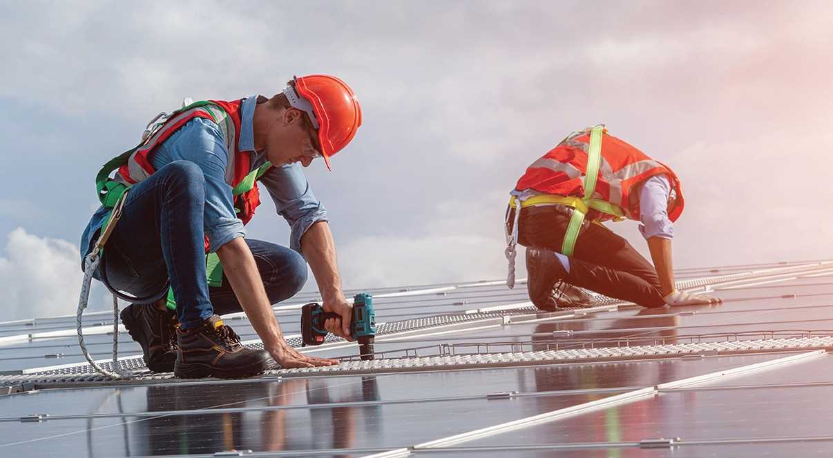 Two individuals in safety gear drill in solar panels on a roof
